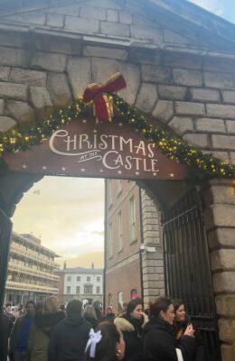 Christmas at the Castle is adorned on a wooden sign above a gate. The Castle bricks are on the exterior of the sign. A lady with a white bow is in the centre front.