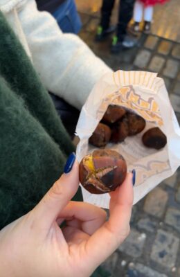 A white hand, with blue artificial nails holds a roasted chestnut at a Christmas Market.