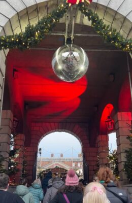 A disco ball hangs from the internal roof of Dublin Castle at the Entrance to the Christmas at the Castle market. There are christmas trees lining the sides of the image.